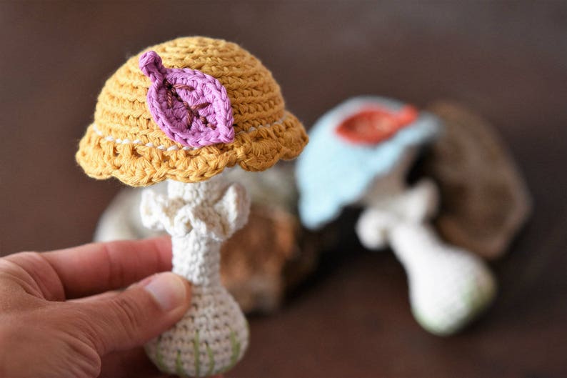 May include: A crocheted mushroom toy with a yellow cap and a pink leaf. The mushroom has a white stem and is made with a soft yarn.
