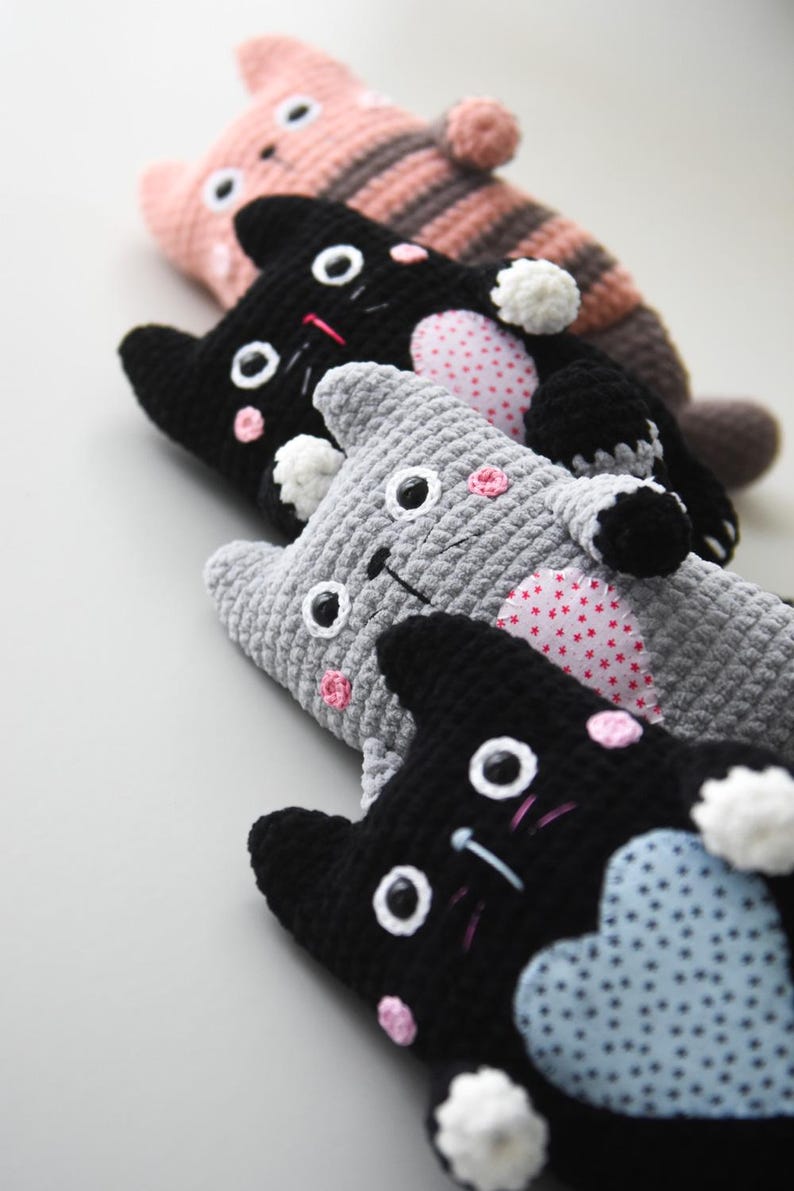 Pode incluir: Uma cole&ccedil;&atilde;o de bichos de pel&uacute;cia de gato de croch&ecirc; feitos &agrave; m&atilde;o em v&aacute;rias cores. Os gatos t&ecirc;m olhos brancos, bochechas rosadas e rostos bordados. Alguns gatos t&ecirc;m cora&ccedil;&otilde;es estampados na barriga, e outros t&ecirc;m corpos listrados ou s&oacute;lidos.