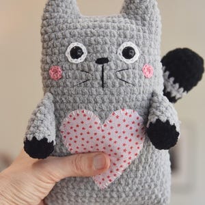 Pode incluir: Um gato de pel&uacute;cia de croch&ecirc; cinza feito &agrave; m&atilde;o com olhos pretos e brancos, bochechas rosa e um cora&ccedil;&atilde;o vermelho com estrelas brancas. O gato tem patas pretas e uma cauda listrada em preto e branco.