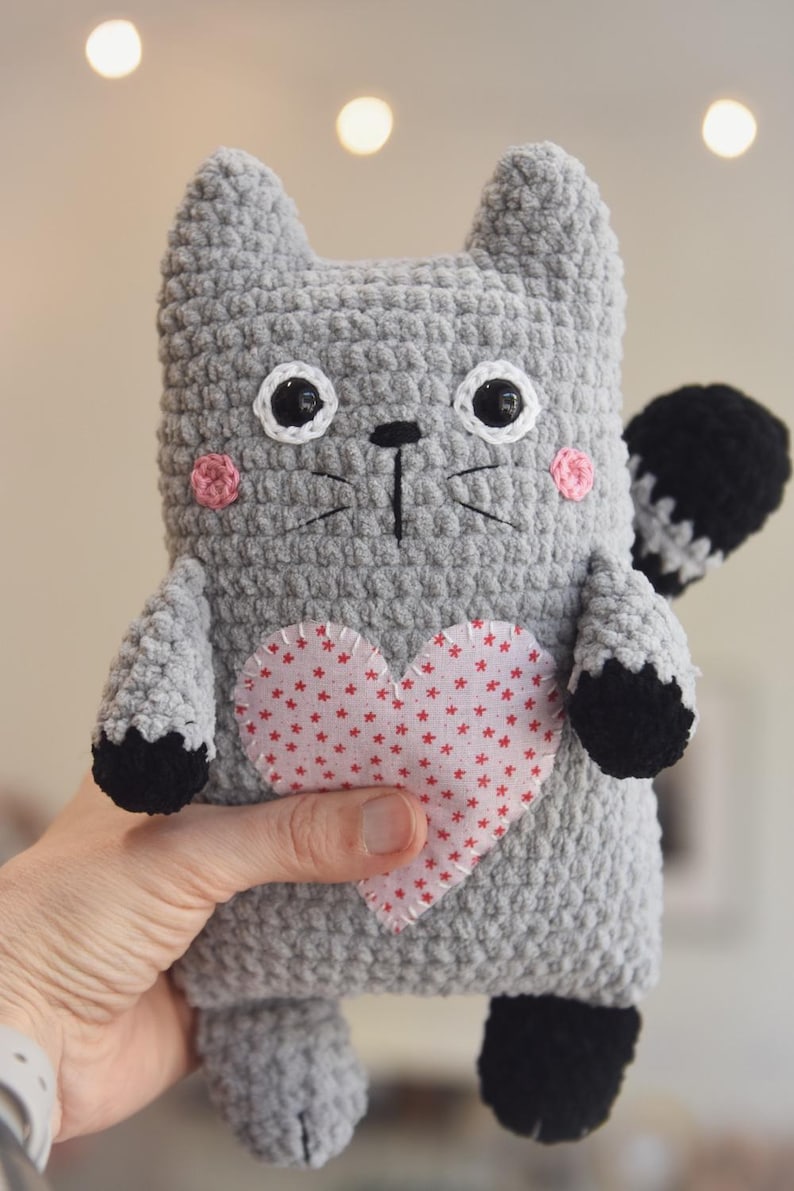 Pode incluir: Um gato de pel&uacute;cia de croch&ecirc; cinza feito &agrave; m&atilde;o com olhos pretos e brancos, bochechas rosa e um cora&ccedil;&atilde;o vermelho com estrelas brancas. O gato tem patas pretas e uma cauda listrada em preto e branco.