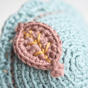 May include: A close-up of a crocheted leaf with a light blue background. The leaf is made of a pink yarn with gold embroidery.