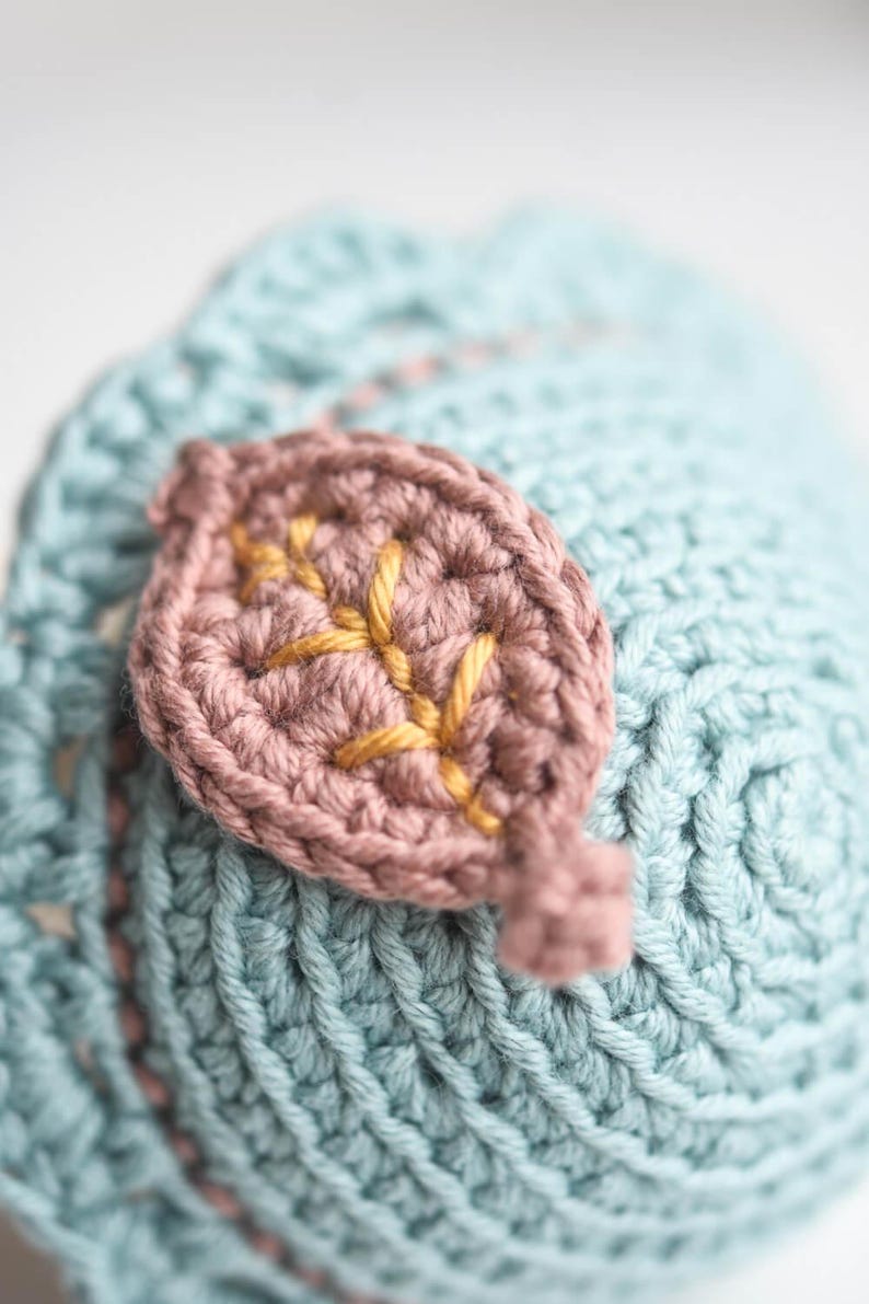 May include: A close-up of a crocheted leaf with a light blue background. The leaf is made of a pink yarn with gold embroidery.