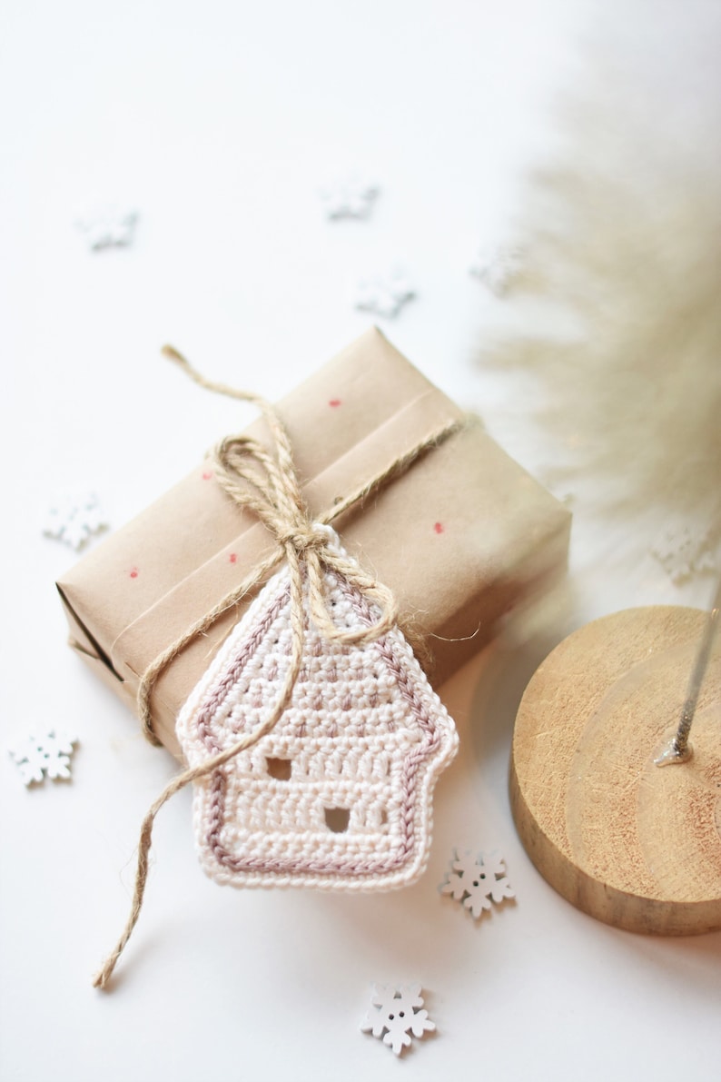 May include: A white crocheted house ornament with a pink trim, tied with twine to a brown gift box wrapped in brown paper with red dots.