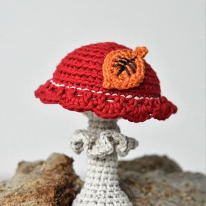 May include: A crocheted mushroom with a red cap and a white stem. The cap has a small orange leaf on top. The mushroom is sitting on a piece of wood.