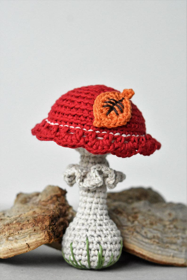 May include: A crocheted mushroom with a red cap and a white stem. The cap has a small orange leaf on top. The mushroom is sitting on a piece of wood.