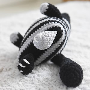 Pode incluir: Um brinquedo de animal de croch&ecirc; feito &agrave; m&atilde;o em listras pretas e cinzas. O brinquedo tem quatro pernas, uma cabe&ccedil;a e uma cauda. O brinquedo est&aacute; sobre uma superf&iacute;cie branca e texturizada.