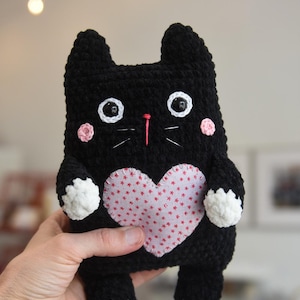 Pode incluir: Um gato de pel&uacute;cia preto feito &agrave; m&atilde;o com um patch em forma de cora&ccedil;&atilde;o rosa no peito. O gato tem detalhes brancos nas patas e bochechas, com bigodes brancos e olhos grandes e redondos. Um bicho de pel&uacute;cia charmoso e fofinho.
