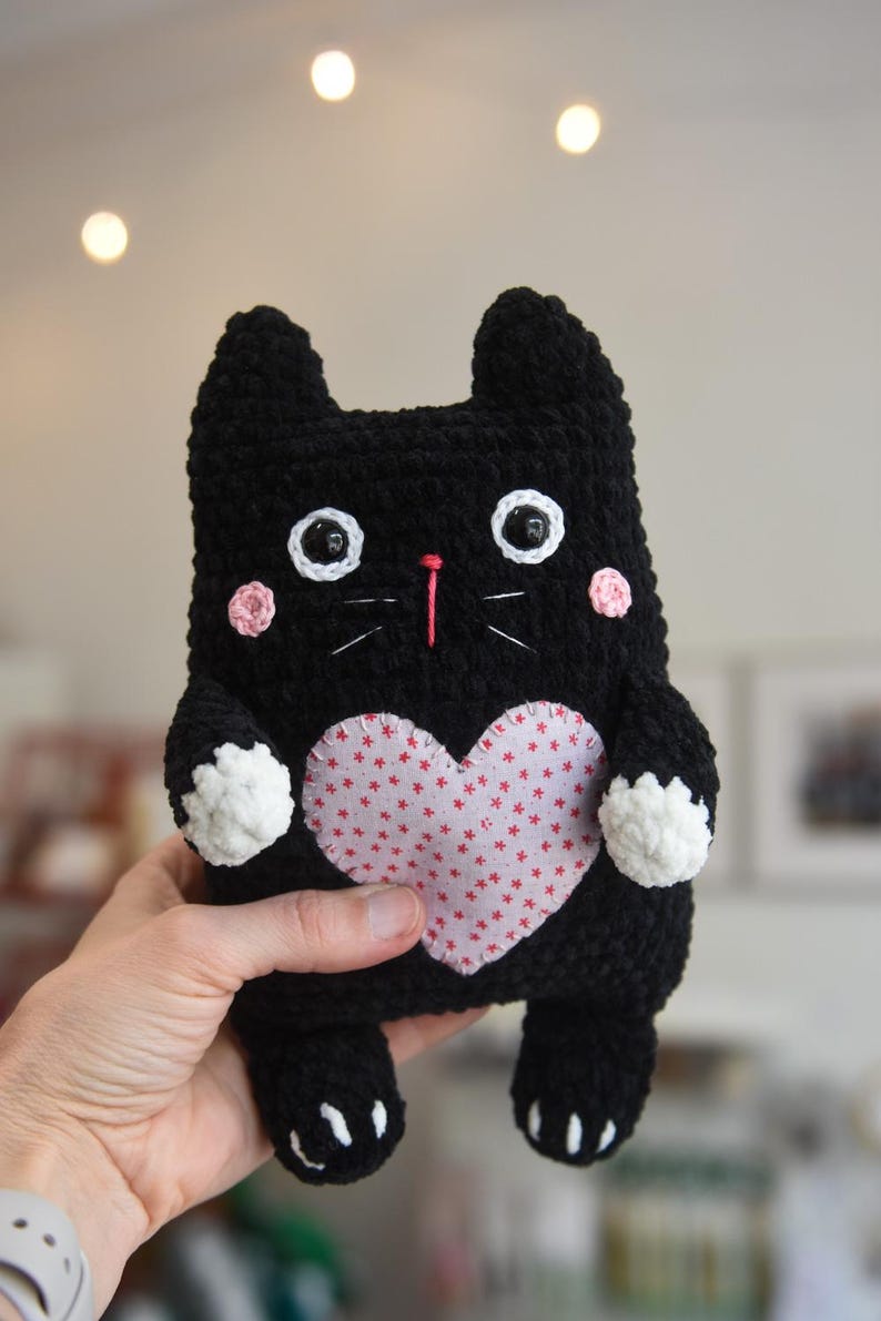 Pode incluir: Um gato de pel&uacute;cia preto feito &agrave; m&atilde;o com um patch em forma de cora&ccedil;&atilde;o rosa no peito. O gato tem detalhes brancos nas patas e bochechas, com bigodes brancos e olhos grandes e redondos. Um bicho de pel&uacute;cia charmoso e fofinho.
