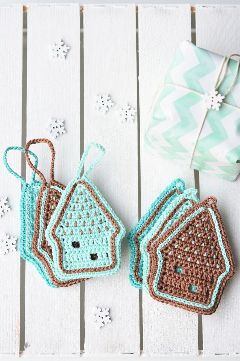 May include: A set of crocheted house ornaments in brown and teal colours. The ornaments are stacked on a white wooden surface with white snowflake decorations.