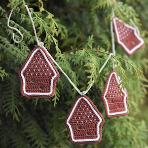 May include: A garland of crocheted gingerbread houses in brown and white hangs on a green evergreen tree branch.