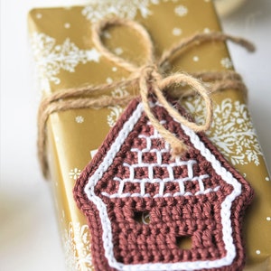May include: A brown crocheted gingerbread house ornament with white trim, tied with twine to a gold gift box with a white snowflake pattern.