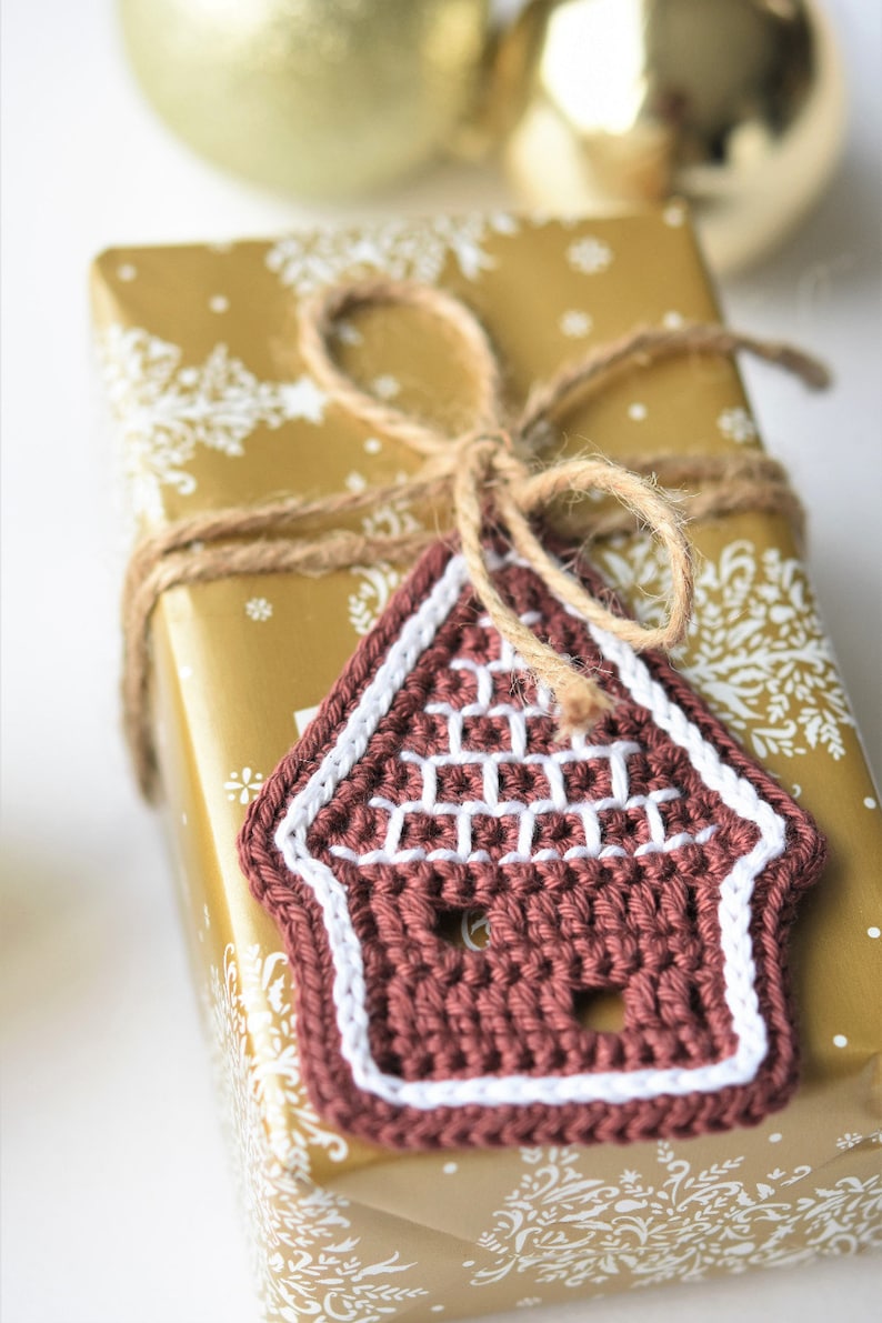May include: A brown crocheted gingerbread house ornament with white trim, tied with twine to a gold gift box with a white snowflake pattern.
