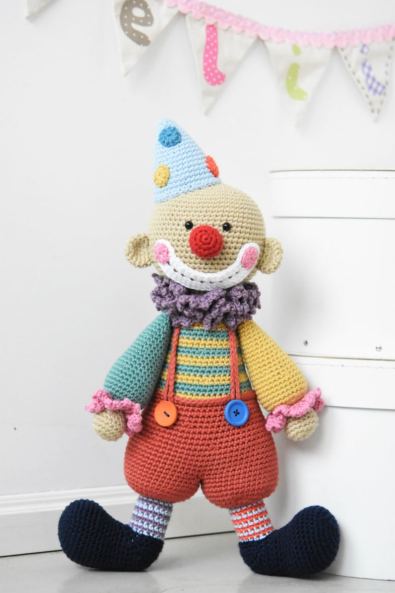 Op de afbeelding: Een gehaakte clownpop met een blauwe en witte hoed, een rode neus en een geel en groen gestreept shirt. De pop draagt een rode broek met knopen en zwarte schoenen.