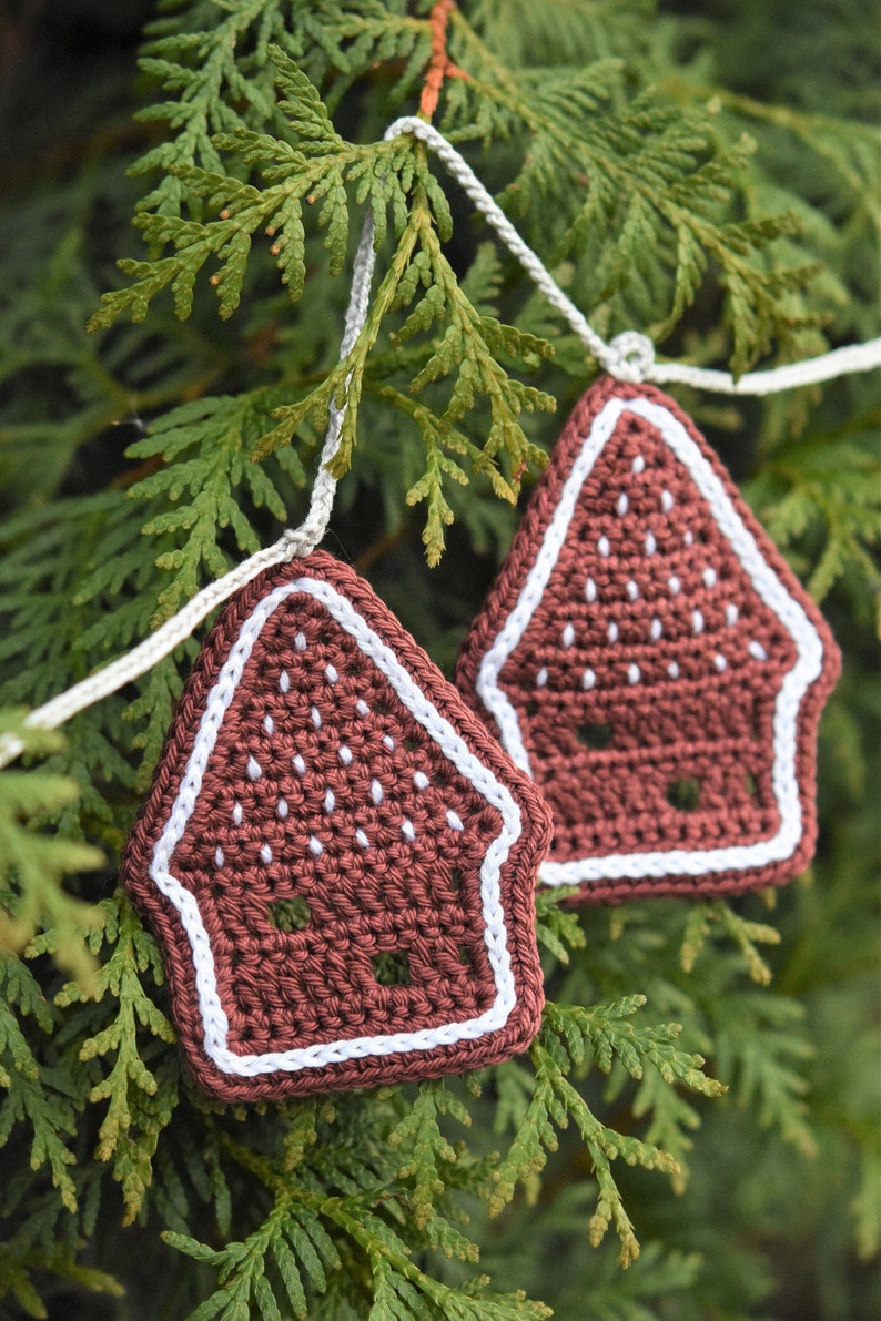 May include: A garland of crocheted gingerbread house decorations in brown and white, hanging from a green evergreen tree branch.