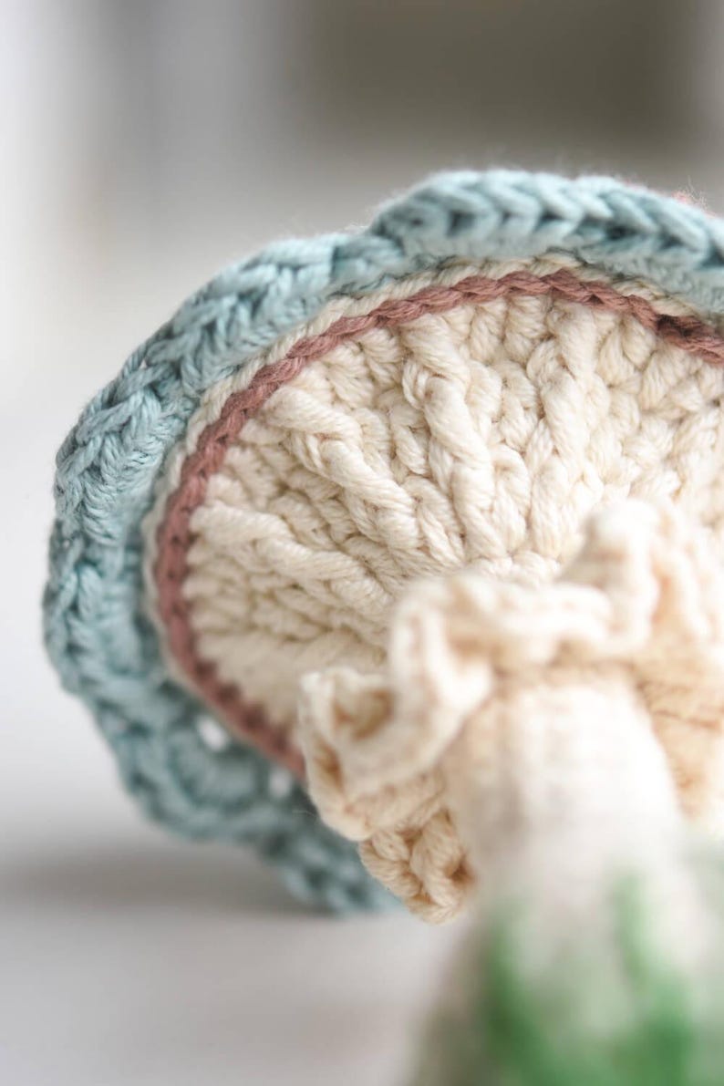 May include: A crocheted mushroom toy with a light blue, pink, and white color scheme. The mushroom has a textured, ribbed top and a ruffled bottom.