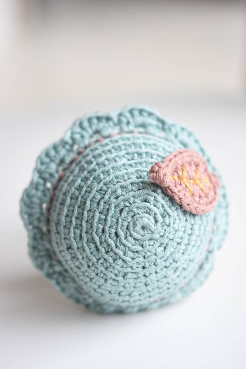 May include: A crocheted ball toy in light blue with a pink leaf detail. The leaf has a yellow embroidered design.