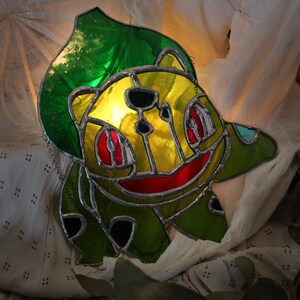 Puede incluir: Atrapasoles de Bulbasaur de vidrieras con una hoja verde, cuerpo amarillo, boca roja y detalles negros. El Bulbasaur está delineado en plata y tiene una cadena para colgar. Una obra de arte única.