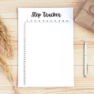 Op de afbeelding: Een witte Step Tracker-pagina met zwarte tekst, een pen en een klein verpakt cadeau op een licht houten oppervlak. De tracker is ontworpen om dagelijkse stappen te monitoren, met markeringen van 1K tot 11K en genummerde dagen van 1 tot 31.