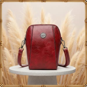 May include: A burgundy leather crossbody bag with a sunburst emblem on the front. The bag has a zip closure, adjustable strap, and is set on a white, round surface. The bag is a stylish accessory.