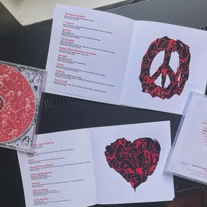 Pu&ograve; includere: Un CD e la sua custodia, insieme a libretti aperti, sono esposti. Il CD e i libretti presentano opere d'arte rosse e nere, tra cui un simbolo di pace e un cuore. Il testo sui libretti elenca i titoli delle canzoni e i crediti.