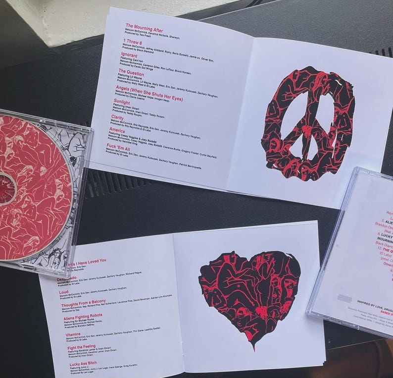 Pu&ograve; includere: Un CD e la sua custodia, insieme a libretti aperti, sono esposti. Il CD e i libretti presentano opere d'arte rosse e nere, tra cui un simbolo di pace e un cuore. Il testo sui libretti elenca i titoli delle canzoni e i crediti.