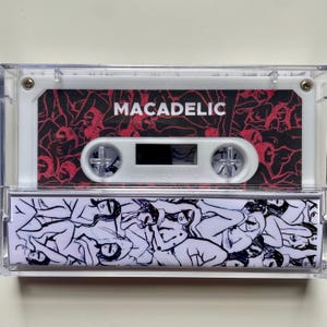 Può includere: Una musicassetta in una custodia di plastica trasparente. L'etichetta della cassetta presenta la parola "MACADELIC" in testo bianco su sfondo rosso e nero. La custodia della cassetta ha un'illustrazione in bianco e nero sul fondo.
