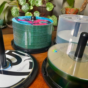 Può includere: Una grande pila di dischi CD-R verdi con una grafica colorata sul disco superiore, accanto a una pila più piccola di dischi CD-R argentati Verbatim e un singolo disco con un design grafico in bianco e nero, montati su perni in plastica nera.
