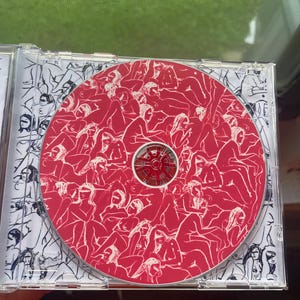 Pu&ograve; includere: Un astuccio per CD con un disco rosso con un motivo di figure. La custodia ha un'illustrazione in bianco e nero sulla copertina interna. Il disco ha un centro rosso e un motivo di figure.