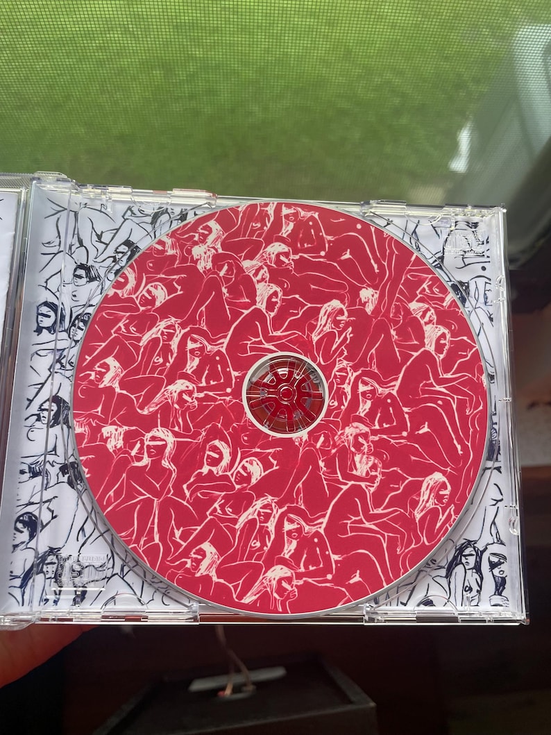 Pu&ograve; includere: Un astuccio per CD con un disco rosso con un motivo di figure. La custodia ha un'illustrazione in bianco e nero sulla copertina interna. Il disco ha un centro rosso e un motivo di figure.