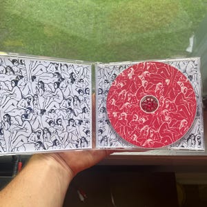 Pu&ograve; includere: Un'apertura di custodia per CD, che rivela un CD rosso con illustrazioni bianche e un'illustrazione in bianco e nero sulla copertina interna. Il CD e la custodia presentano disegni a tratto di figure. La custodia &egrave; tenuta contro uno sfondo verde.