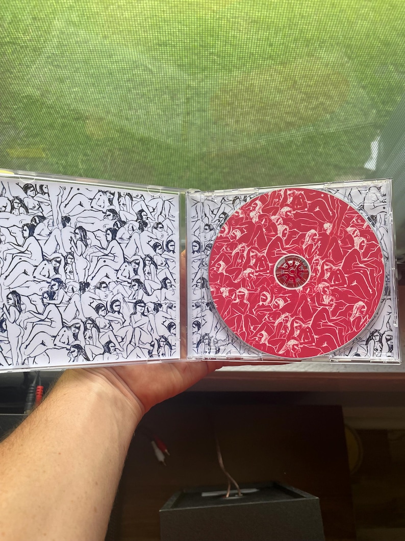 Pu&ograve; includere: Un'apertura di custodia per CD, che rivela un CD rosso con illustrazioni bianche e un'illustrazione in bianco e nero sulla copertina interna. Il CD e la custodia presentano disegni a tratto di figure. La custodia &egrave; tenuta contro uno sfondo verde.