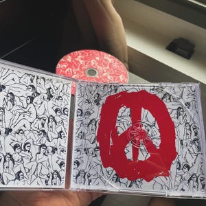 Pu&ograve; includere: Un'apertura di un cofanetto CD, che rivela un CD e il suo contenuto. Il cofanetto presenta un simbolo della pace rosso su sfondo bianco con disegni di figure in nero. Il CD &egrave; rosso con un motivo bianco.