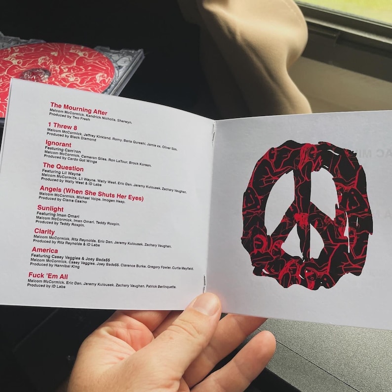 Pu&ograve; includere: Un libretto di CD aperto che mostra titoli di canzoni e crediti. Un simbolo della pace rosso e nero &egrave; sulla pagina di destra. Il libretto &egrave; tenuto in mano, con una custodia per CD visibile sullo sfondo.