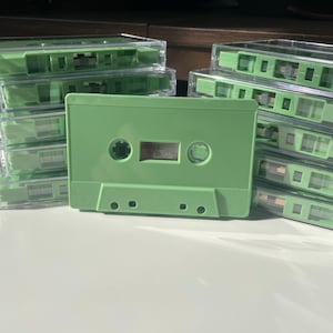 May include: A stack of blank audio cassette tapes with light green plastic shells. The cassettes feature a rectangular design with two circular spools and a central window. The shells are solid light green, and the tapes are housed in clear plastic cases.