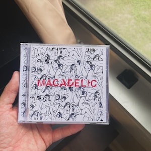 Op de afbeelding: Een cd-hoesje met een zwart-wit illustratie van talrijke figuren. Het woord "MACADELIC" is in het rood op de voorkant gedrukt. De hoes wordt in een hand gehouden, met een raam en een blik naar buiten op de achtergrond.