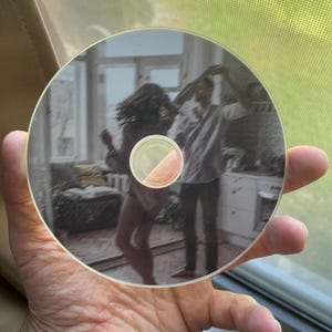 May include: A clear CD with a printed image of two people dancing in a room. The image is in muted tones, with the figures appearing blurred. The CD is held in a hand, with a window and screen visible in the background.