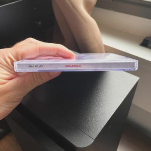 Pu&ograve; includere: Una custodia per CD in plastica trasparente con il testo "MAC MILLER" e "MACADELIC" in rosso. La custodia &egrave; tenuta in mano da una persona, con un altoparlante nero sullo sfondo.
