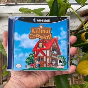 Può includere: Custodia del gioco Nintendo GameCube per "Animal Crossing". La copertina presenta una casa a cartoni animati con tetto rosso, cielo blu e la scritta "Welcome to Animal Crossing". Il gioco è classificato "E" per Tutti.