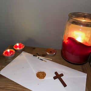 Op de afbeelding: Een brandende rode kaars in een glazen pot, omringd door drie brandende waxinelichtjes. Een witte envelop met een waszegel en een houten kruis liggen op een houten oppervlak. Twee gebruikte lucifers zijn ook aanwezig.