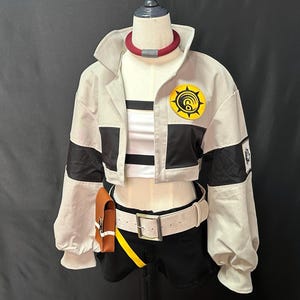 Może przedstawiać: Strój cosplay na manekinie. Strój zawiera jasno szarą kurtkę z czarnymi akcentami, biały top z czarnymi paskami i czarne szorty. Brązowa torebka wisi na białym pasku. W zestawie znajduje się również czerwono-szary naszyjnik.