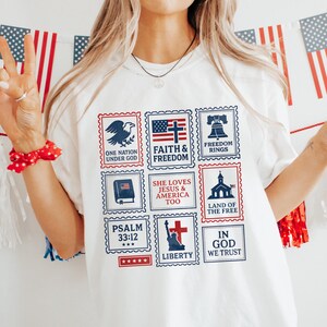以下が含まれることがあります： 白いTシャツ。愛国的な切手デザインで、ワシ、アメリカ国旗、「Faith & Freedom」の文字が特徴です。シャツには「She Loves Jesus & America Too」と「In God We Trust」の文字も含まれています。