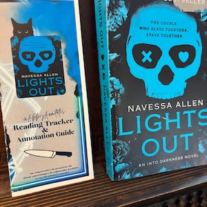 May include: Two books and a reading guide are displayed on a wooden shelf. The book on the right is titled "LIGHTS OUT" by Navessa Allen, with a skull graphic. The reading guide has a black cat graphic and the text "Reading Tracker & Annotation Guide."