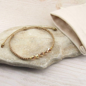 Peut inclure: Un bracelet en cordon beige orné de quatre perles en cristal à facettes de couleur or clair disposées en ligne. Le bracelet présente une structure en cordon tressé fin avec de petites perles métalliques aux extrémités et une pochette en tissu écru.