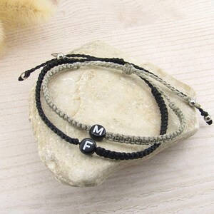 Könnte beinhalten: Zwei verstellbare Kordelarmbänder, eines in Schwarz und eines in Hellgrau, mit jeweils einer runden schwarzen Perle mit weißem Buchstaben. Das schwarze Armband zeigt den Buchstaben F, das hellgraue Armband den Buchstaben M, mit Silberperlen.