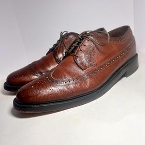 May include: A pair of brown leather dress shoes with a textured finish and decorative perforations. The shoes have a classic wingtip design, dark brown laces, and black soles. The shoes are displayed on a white surface.