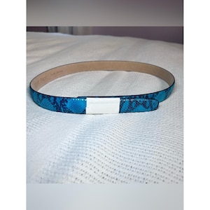 May include: A thin belt featuring a vibrant blue and black snakeskin pattern. The belt has a smooth, light-colored interior lining and a rectangular white protective paper strip wrapped around the front section of the strap.