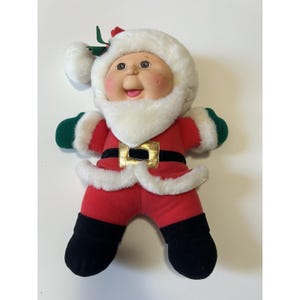 May include: A plush Santa Claus doll with a round face, rosy cheeks, and a white beard. The doll wears a red suit with white trim, a black belt with a gold buckle, and black boots. Green mittens and a white fur-trimmed hat complete the festive look.