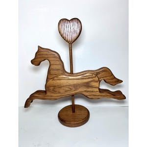May include: Wooden horse sculpture with a heart-shaped accent. The horse is carved in a running pose and mounted on a round base. The heart is positioned above the horse, connected by a wooden rod. The sculpture is made of a light brown wood.