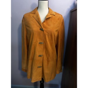 May include: Mustard-colored suede jacket with a collared neckline and long sleeves. The jacket features three wooden toggle closures down the front. The jacket is displayed on a mannequin.