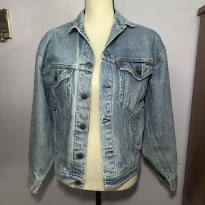 May include: Light blue denim jacket with a classic design. Features a button-up front, two chest pockets, and a red logo tag. The jacket is displayed on a mannequin.